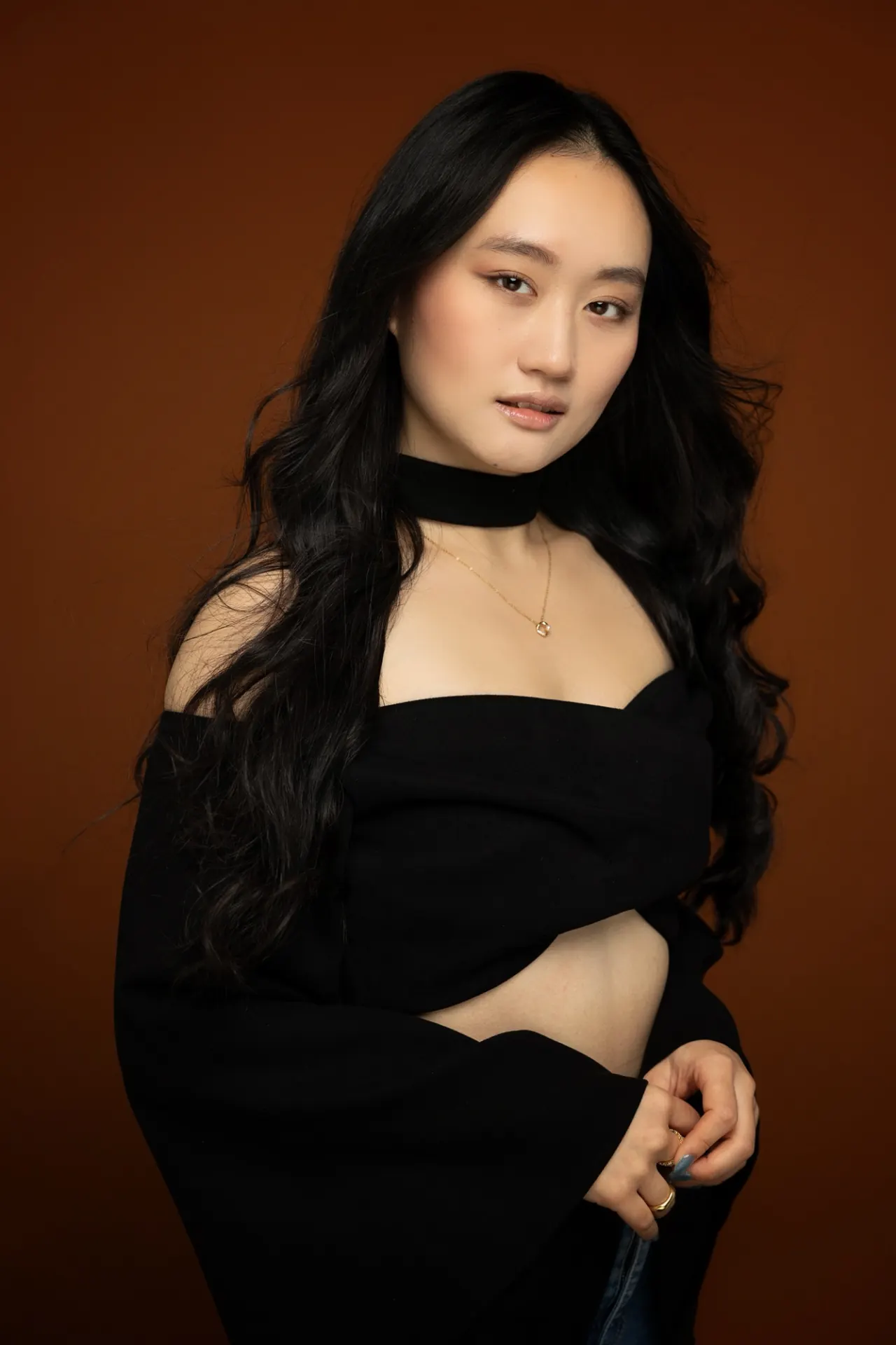 AJ Ho in a black off-shoulder dress against a rust-coloured backdrop