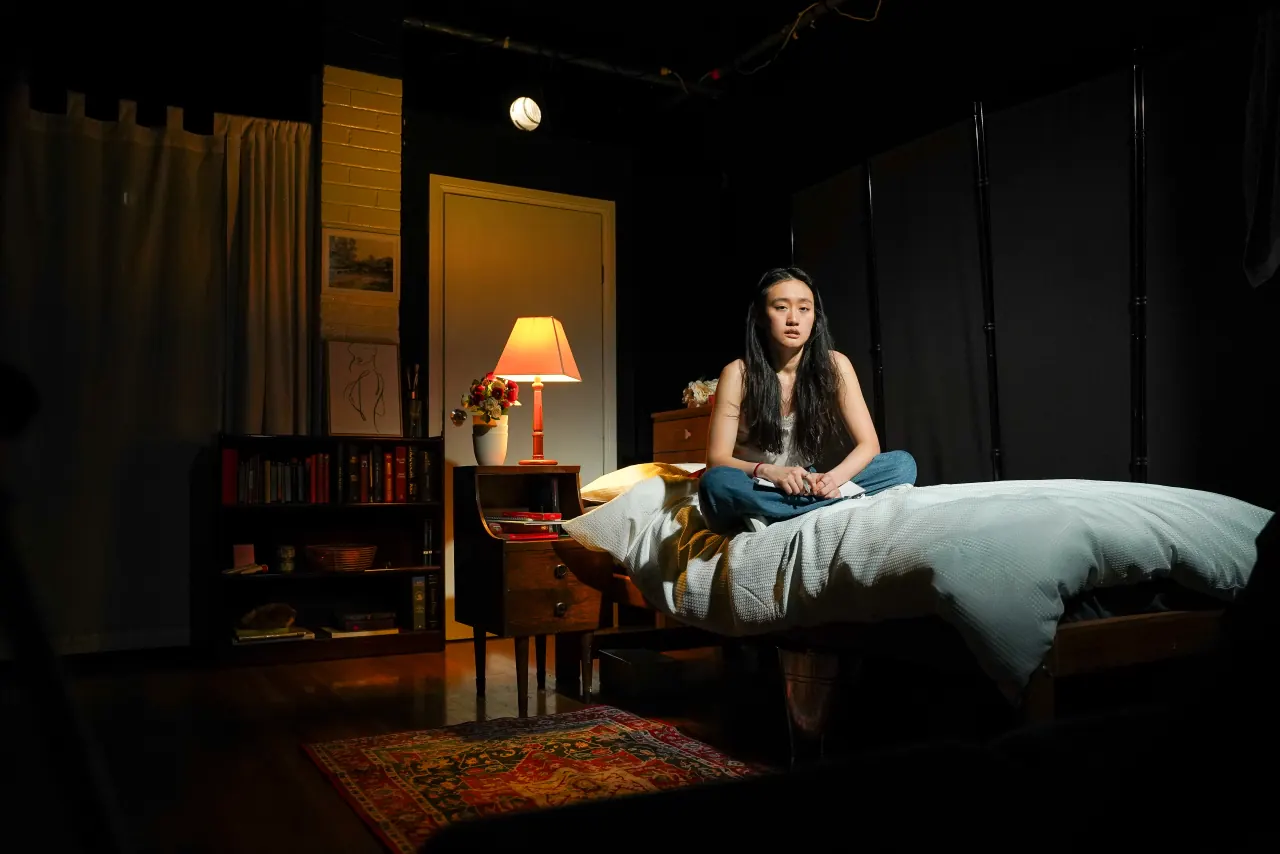 AJ Ho as Miranda Grey sitting alone cross-legged on the bed of the cellar room, lit by a single lamp