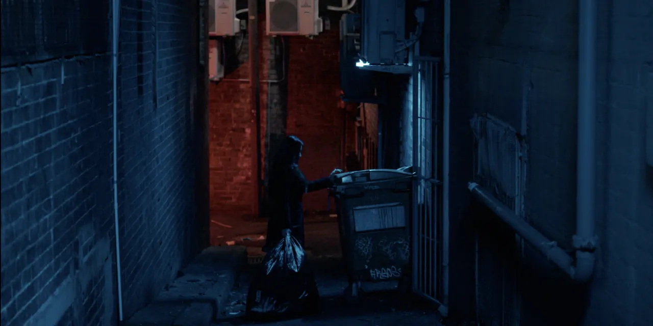 Still from God I&rsquo;m Starved: a figure alone in a blue-lit alley beside a dumpster